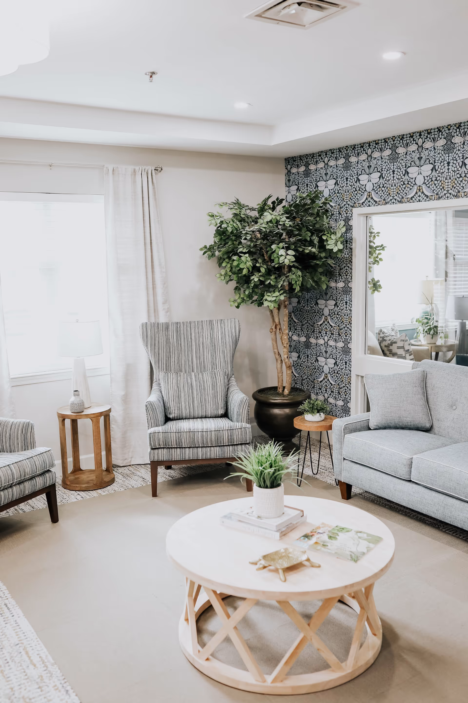 A bright living room with upholstered armchairs and a sofa, a round wooden coffee table with a potted plant, and a large potted tree against patterned wallpaper.