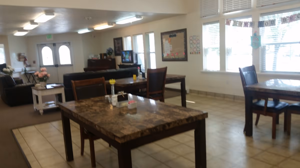 Communal dining and activity room with several tables and chairs, windows along one wall and a seating area in the background.
