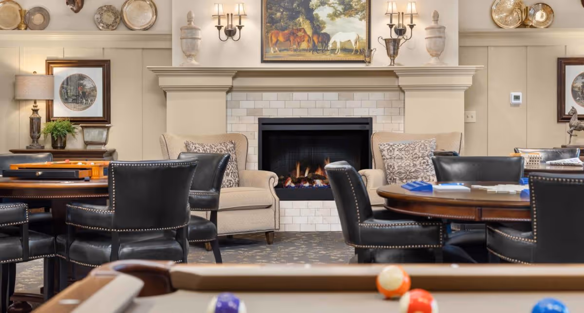 Cozy common room with a fireplace, upholstered chairs, card tables, and a pool table in the foreground.
