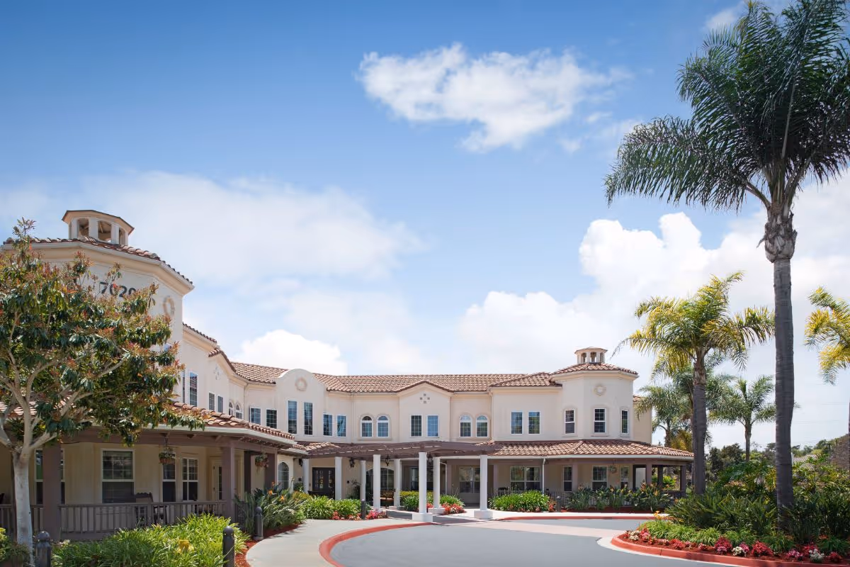 Front exterior of a Mediterranean-style senior living building with a covered portico, palm trees, and a circular driveway under a blue sky.
