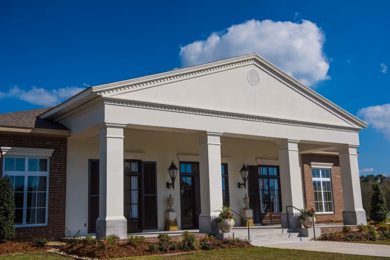 Front exterior view of a single-story building with white columns, large windows, and a triangular pediment under a blue sky with some clouds. There are plants and a small garden in front of the building.