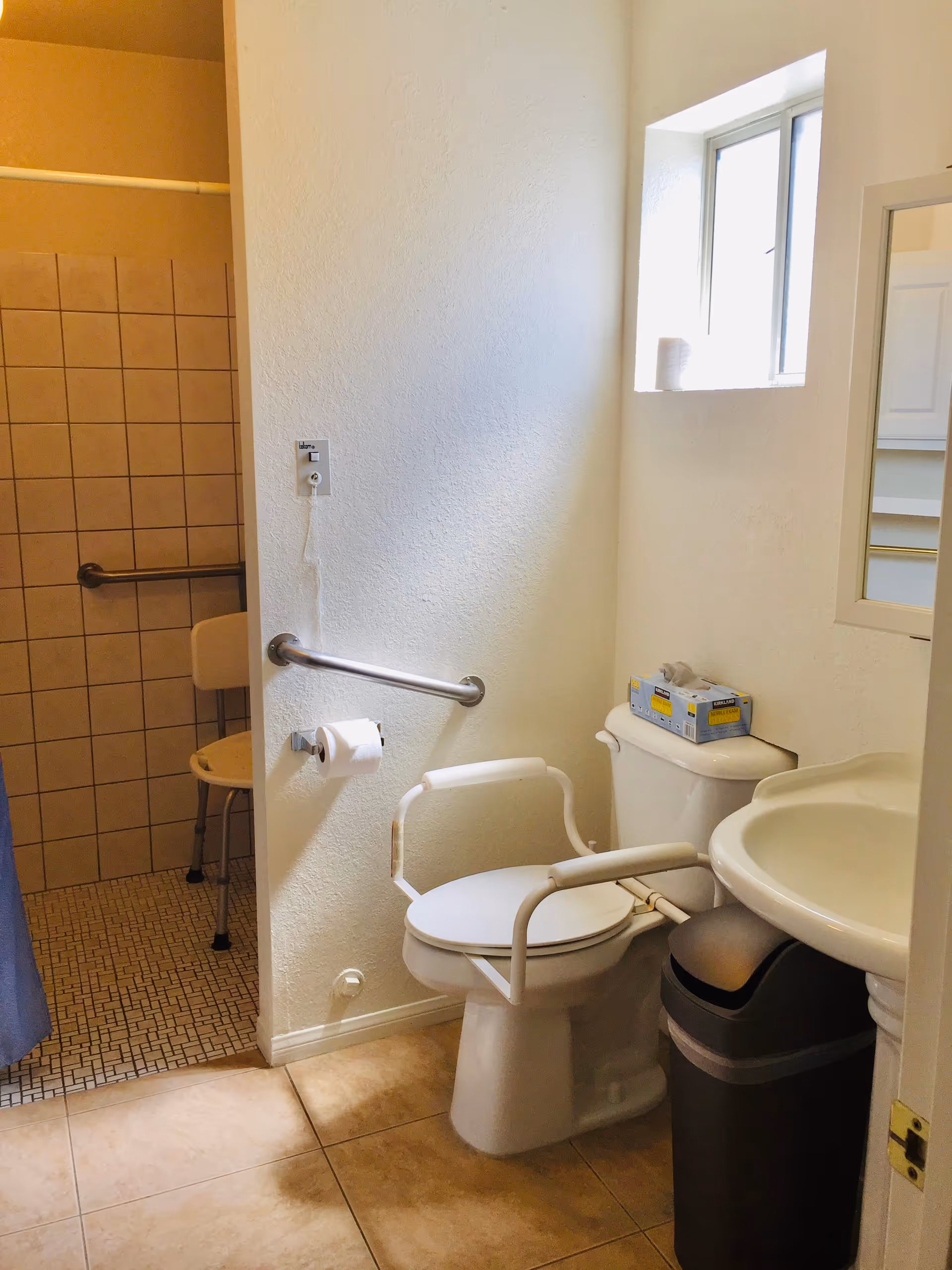 A bathroom with a toilet equipped with white safety rails, a wall-mounted grab bar, and a toilet paper holder. There is a window above the toilet with a roll of toilet paper on the sill. To the right is a white sink with a mirror above it and a trash can below. In the background, there is a tiled shower area with a shower chair and grab bars.