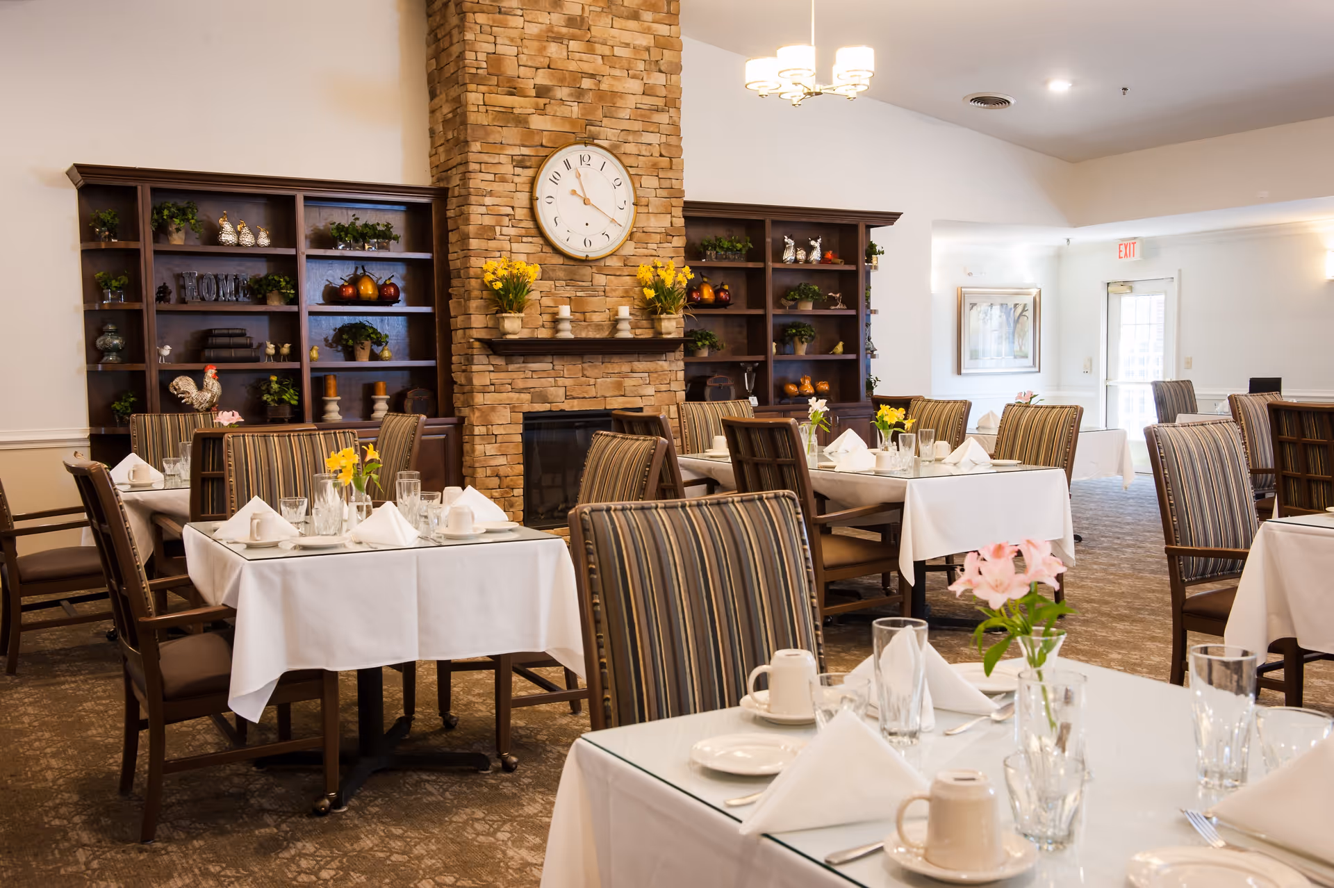 A dining room in a senior living facility with tables covered in white tablecloths, set with glasses, cups, plates, and napkins. The room features striped upholstered chairs, a stone fireplace with a clock above it, and wooden shelves decorated with plants and ornaments. The space is well-lit with ceiling lights and natural light from a door in the background.