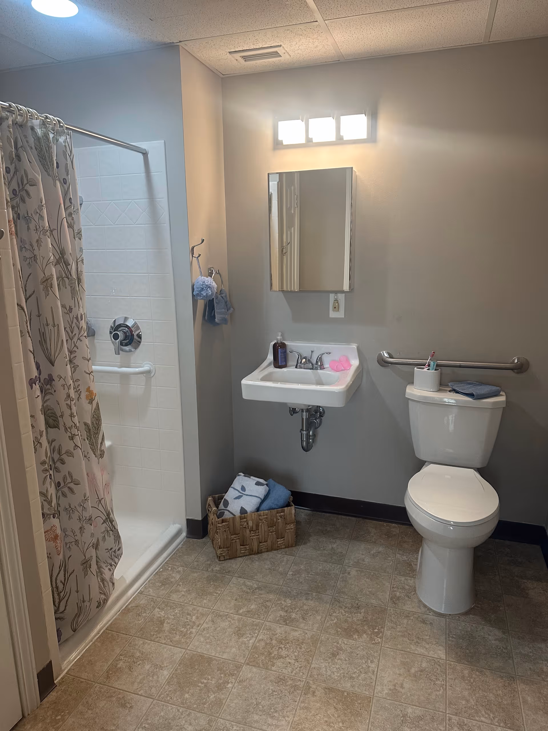 A clean and accessible bathroom featuring a shower with a floral-patterned curtain, a wall-mounted sink with a mirror above it, and a toilet with a grab bar on the wall beside it. There is a basket with folded towels on the floor under the sink, and a soap dispenser and pink sponge on the sink. The walls are painted light gray and the floor has beige tiles.
