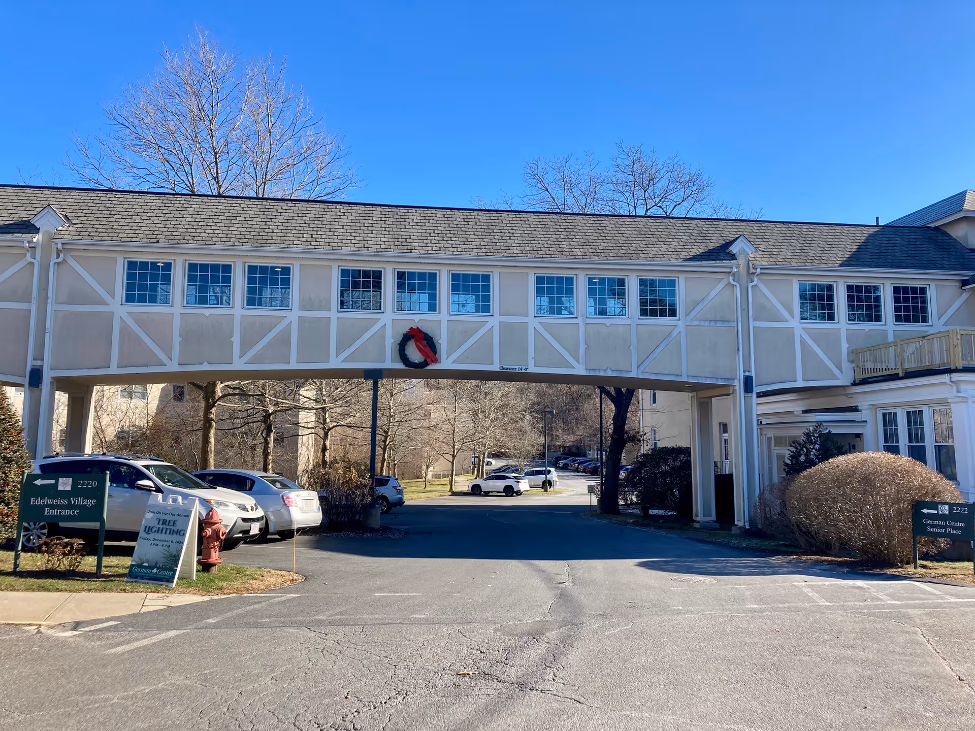 Exterior view of a senior living facility entrance with a covered walkway connecting two buildings. There are parked cars on the left side, a fire hydrant, and signs indicating directions to Edelweiss Village Entrance and German Centre Senior Place. A holiday wreath is hanging on the covered walkway. Trees without leaves are visible in the background under a clear blue sky.
