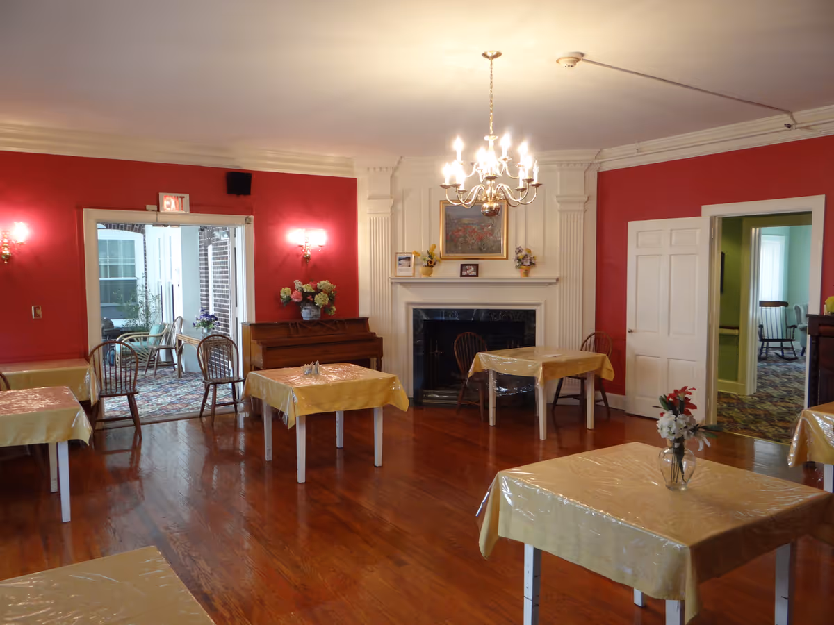 A cozy dining room with red walls and wooden floors featuring several small tables covered with yellow tablecloths and plastic covers. A chandelier hangs from the ceiling above a white fireplace adorned with framed pictures and flower arrangements. There is a piano against one wall and an exit door leading to a sunroom with additional seating. Another doorway opens to a room with green walls and a rocking chair.