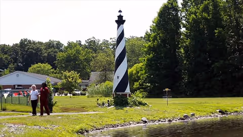 Two people walking on a path near a pond with a black and white striped lighthouse structure and surrounded by green trees and grass.