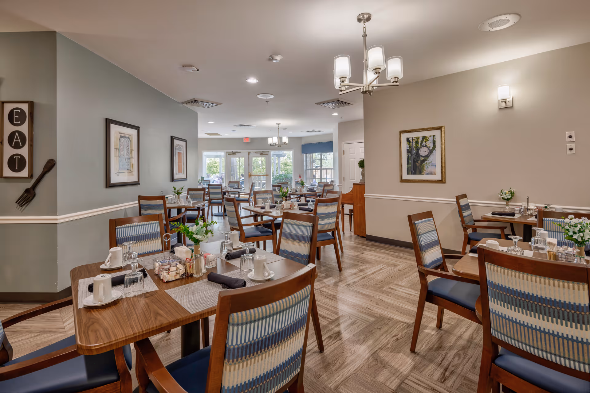 A bright and neatly arranged dining room with multiple wooden tables and chairs featuring blue and beige patterned upholstery. Each table is set with cups, glasses, napkins, and small flower arrangements. The room has light-colored walls with framed artwork and decorative wall hangings, including a sign that says 'EAT' with a large fork. Ceiling lights and wall sconces illuminate the space, and large windows at the far end let in natural light.