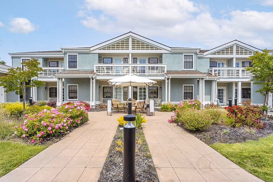 Exterior view of a two-story senior living facility building with light blue walls, white trim, and multiple windows. The building features balconies on the upper floor and a patio area with outdoor seating and an umbrella. The foreground shows a paved walkway flanked by landscaped flower beds with pink and red flowers and green shrubs under a partly cloudy sky.