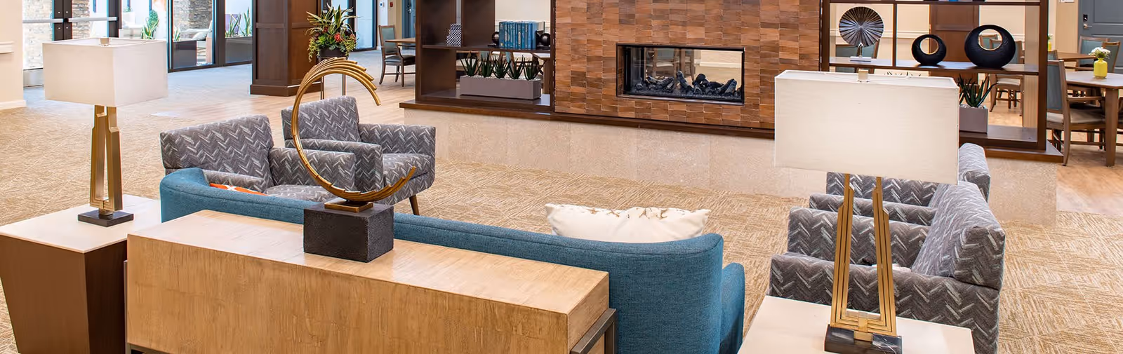 A spacious and well-lit living room area in a senior living facility featuring modern furniture including a blue sofa, patterned armchairs, two table lamps with rectangular shades, a decorative sculpture on a wooden console table, and a built-in fireplace with a stone surround. The background shows a dining area with tables and chairs and large windows letting in natural light.