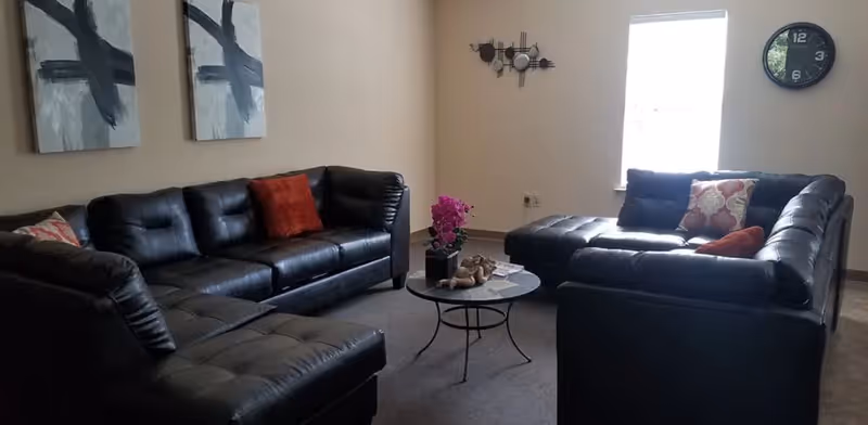 A living room with two black leather sectional sofas arranged around a small round coffee table with a pink orchid and decorative items. The walls are beige with two abstract paintings on one side and a clock and wall decor near a window letting in natural light.