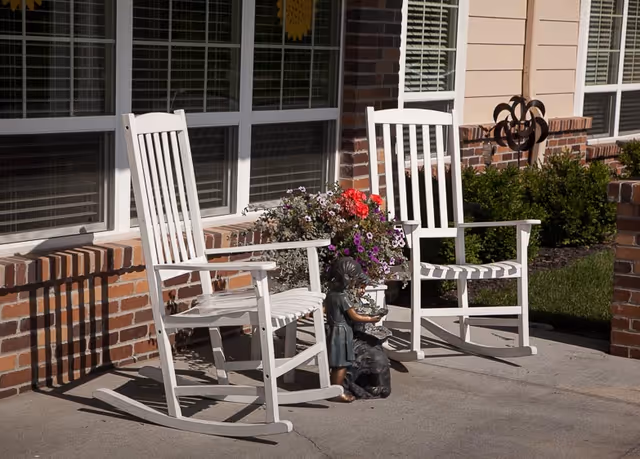 Two white wooden rocking chairs on a concrete patio outside a brick and siding building. Between the chairs is a flower pot with colorful flowers and a small statue of a girl and a dog. There are windows with blinds behind the chairs and some green bushes in the background.