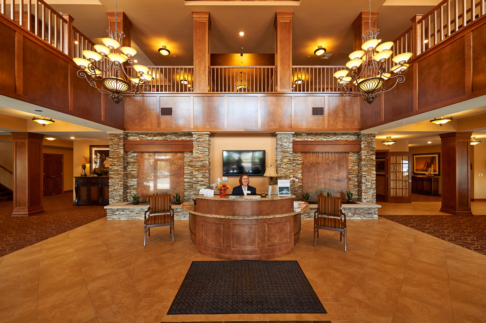 Spacious senior living facility lobby with a wooden reception desk at the center, a receptionist smiling behind the desk, two striped chairs in front, stone accent walls with water features, warm lighting from chandeliers, and a second-floor balcony overlooking the area.