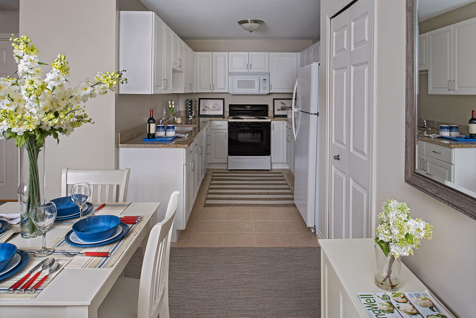 A bright and clean kitchen with white cabinets, a white refrigerator, stove, and microwave. A dining table set with blue plates, wine glasses, and red-handled cutlery is visible in the foreground. There are two striped rugs on the floor, a vase with white and green flowers on the dining table, and another vase with white flowers on a small cabinet with a magazine. A large mirror hangs on the wall above the cabinet.