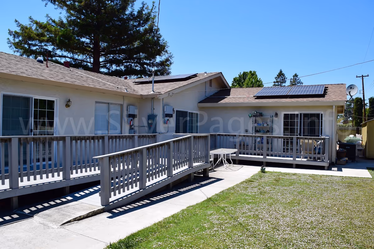 Exterior view of a single-story residential building with a wooden wheelchair ramp and deck, solar panels on the roof, and a small lawn.