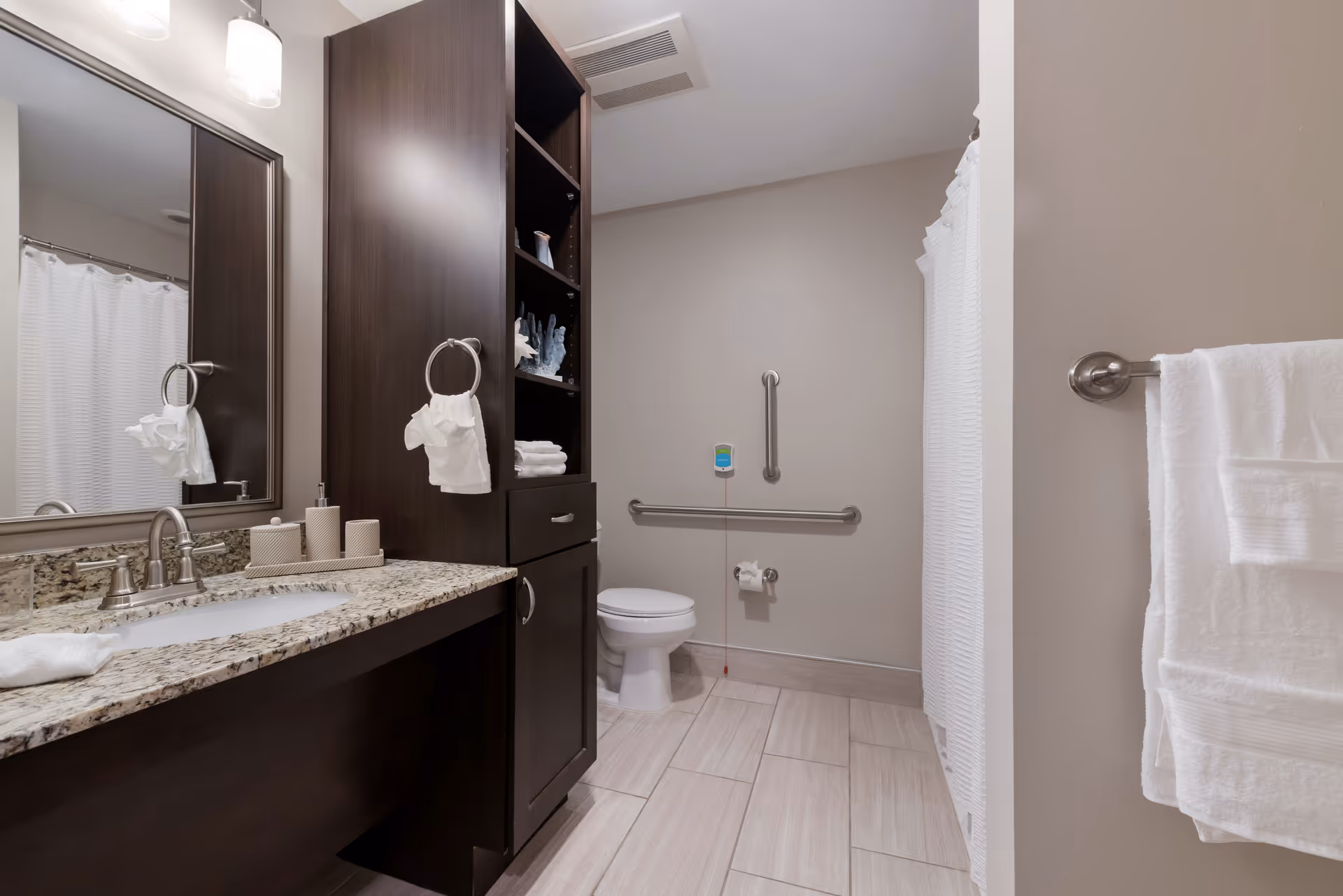 A clean and modern bathroom with a granite countertop sink, dark wood cabinetry, a large mirror, a toilet with grab bars, and a shower with a white curtain. White towels hang on towel rings and racks.