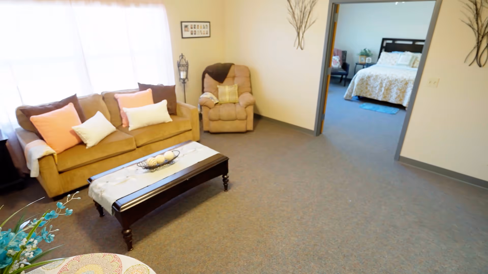 A cozy living room with a beige sofa adorned with pink and white pillows, a matching armchair, and a dark wooden coffee table with a decorative runner and centerpiece. The room has a large window with sheer white curtains allowing natural light to fill the space. In the background, an open doorway leads to a bedroom with a bed covered in a light-colored quilt and a small chair near the window.
