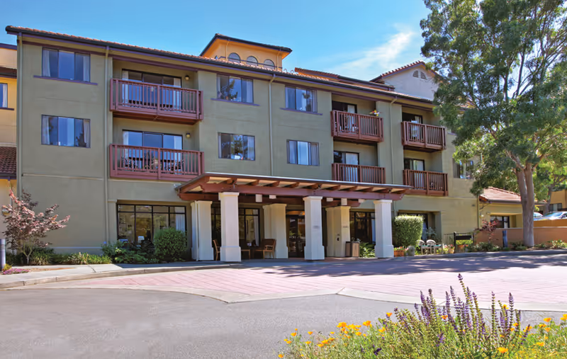 Front exterior of a three-story senior living building with a covered entrance, balconies, and landscaped grounds.