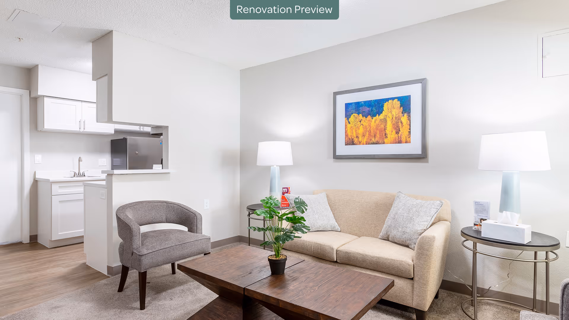 A bright and modern living room with a beige sofa, two gray pillows, a gray armchair, a wooden coffee table with a small potted plant, and two side tables each with a lamp. A colorful framed artwork hangs on the wall above the sofa. The room opens into a small kitchen area with white cabinets and a stainless steel refrigerator. The floor is a light wood laminate, and the walls are painted white. A label at the top center reads 'Renovation Preview'.
