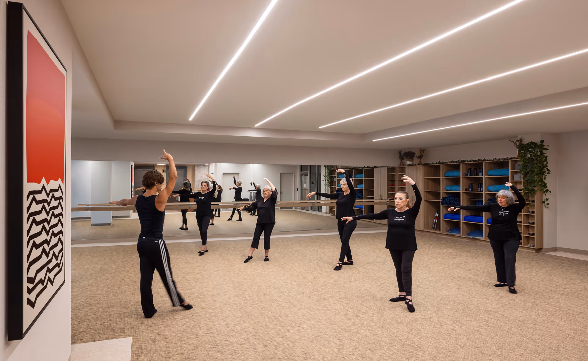 A group of five older adults participating in a dance or exercise class in a spacious, well-lit studio with beige carpet, large mirrors on one wall, and shelves with blue exercise mats and equipment. The instructor is leading the class, and all participants are raising one arm gracefully.