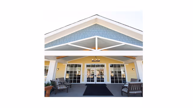 Entrance of a building with a covered porch featuring a peaked roof with blue shingles and white trim. There are glass double doors flanked by large windows, with outdoor seating including cushioned chairs on either side of the entrance. A chandelier hangs from the ceiling of the porch.