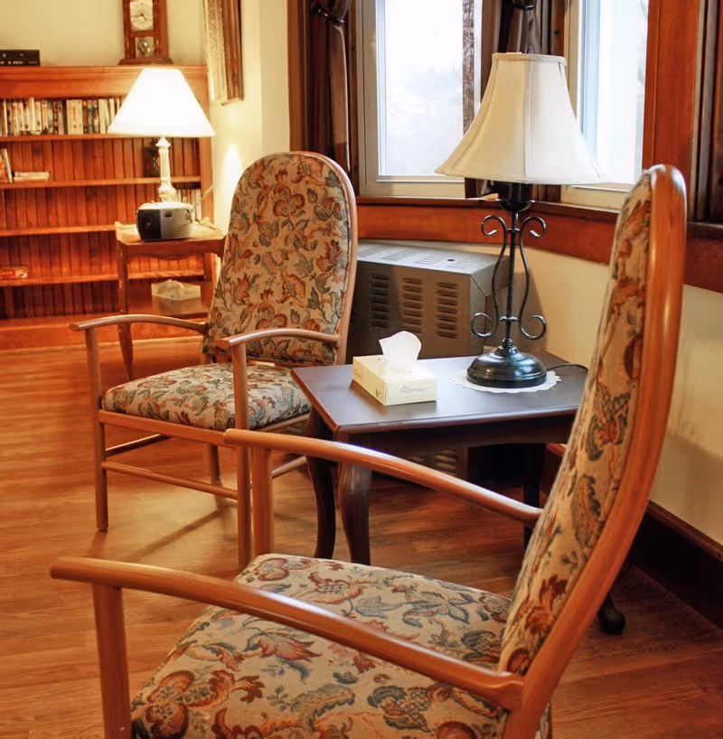 A cozy sitting area with two floral upholstered wooden armchairs facing each other across a small wooden table. On the table is a box of tissues and a decorative lamp with a white lampshade. In the background, there is a wooden bookshelf filled with books and a small radio, with warm lighting creating a comfortable atmosphere.