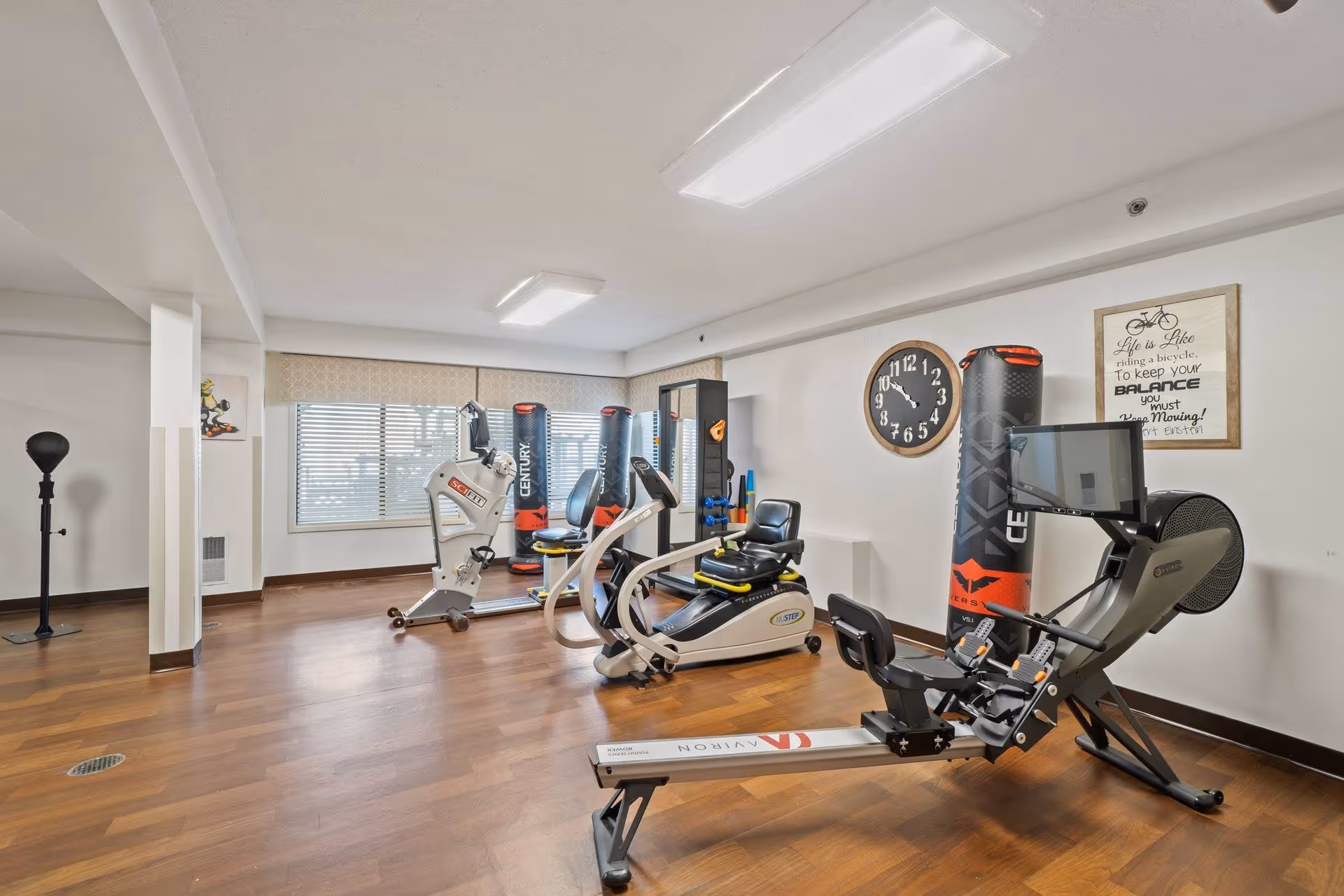 A fitness room with various exercise equipment including a rowing machine, stationary bikes, and punching bags. The room has wooden flooring, white walls, a large window with blinds, a wall clock, and a framed motivational quote about balance and movement.