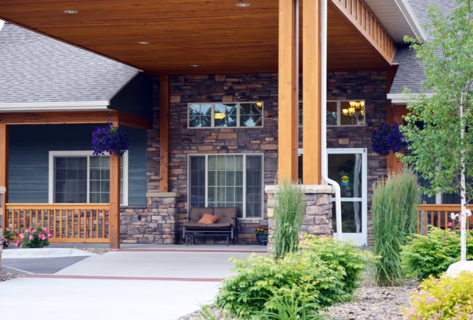 Entrance of Timber Creek Village Assisted Living of Havre showing a covered porch with wooden beams, stone walls, a bench with cushions, hanging flower baskets, and surrounding greenery including bushes and a tree.