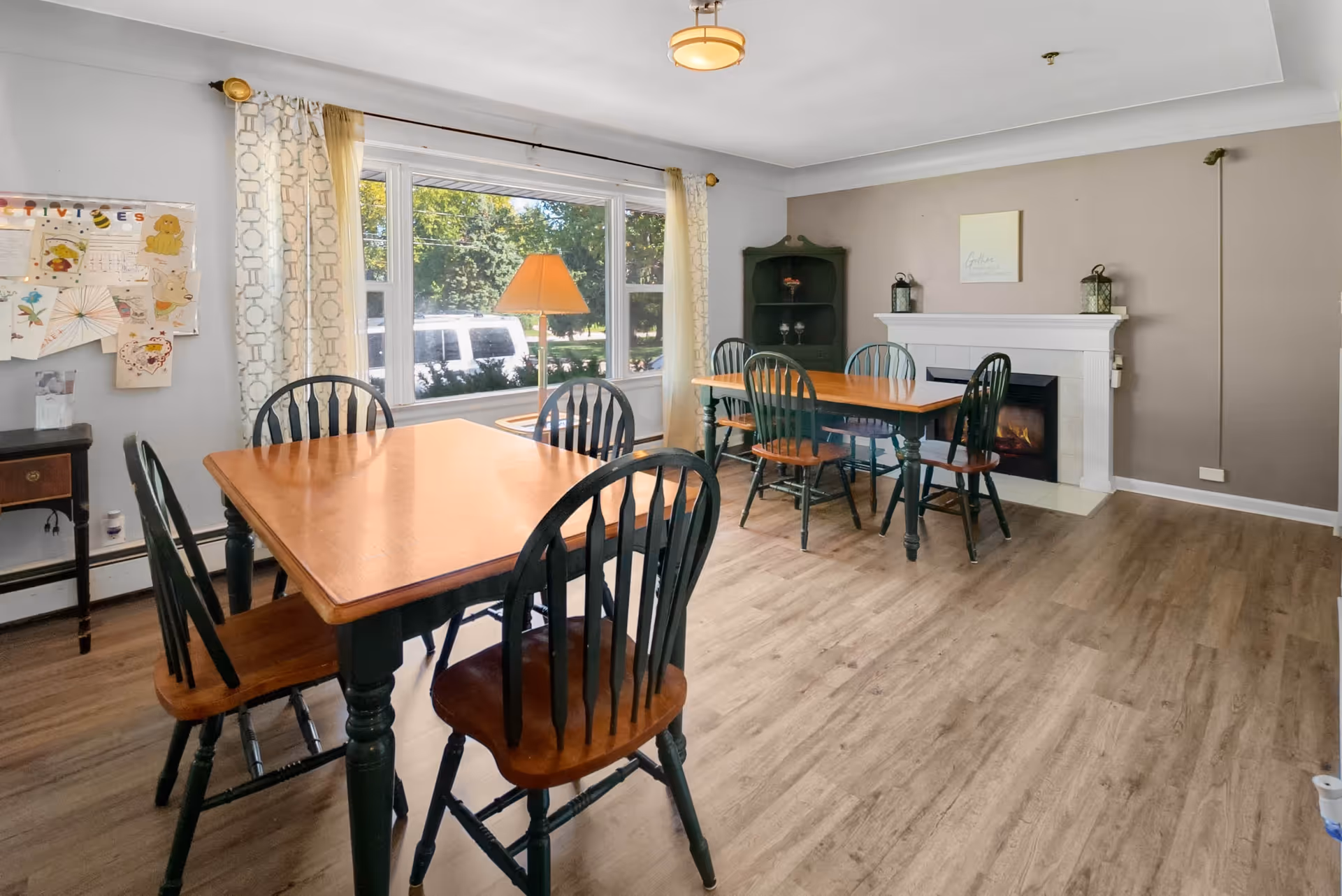 A cozy dining room with two wooden tables and eight matching chairs. The room features a large window with patterned curtains letting in natural light, a floor lamp, a white fireplace with decorative lanterns on the mantel, and a corner cabinet. The floor is wood, and the walls are painted in neutral tones.