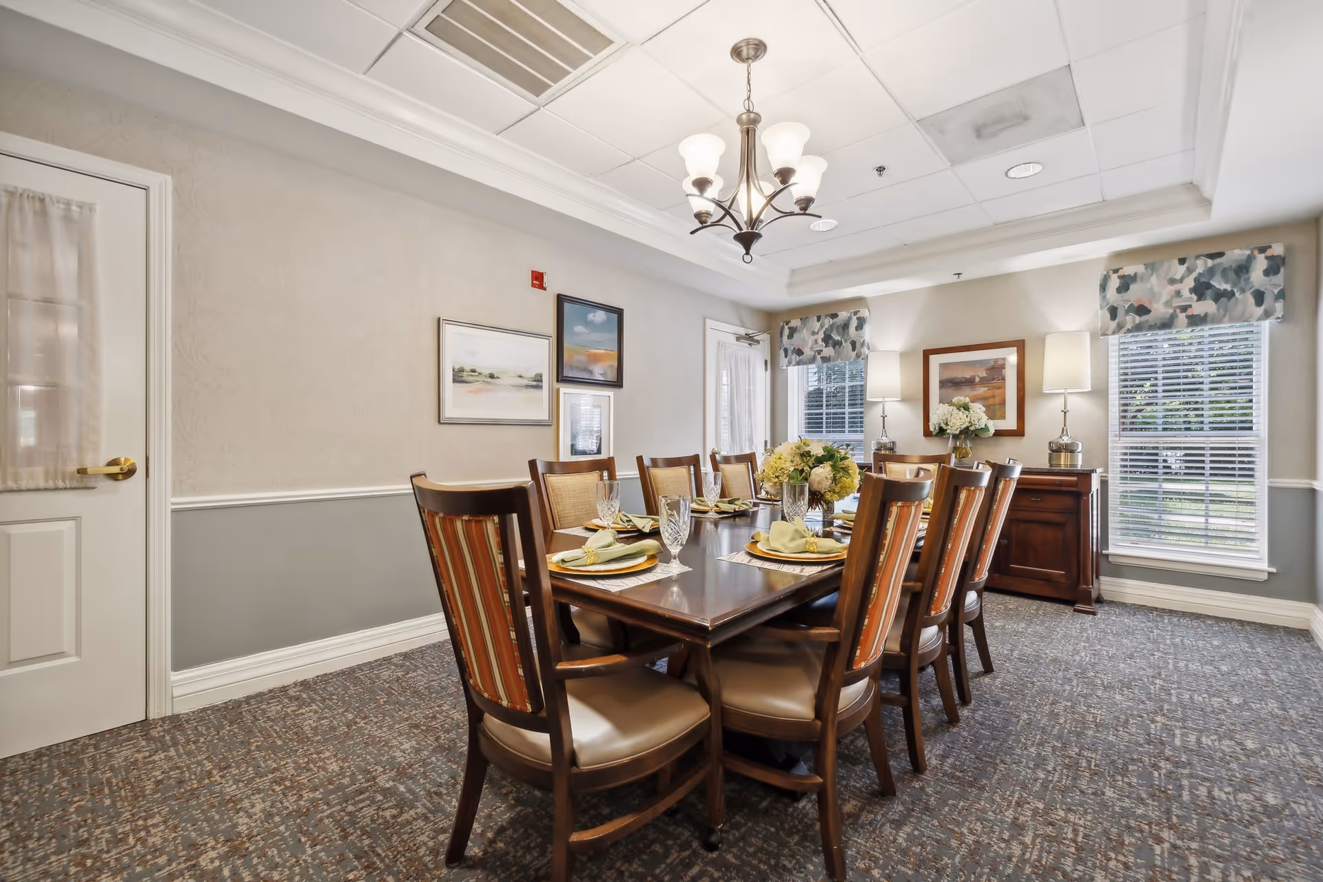 A formal dining room with a long wooden table set for eight, chandelier overhead, artwork on the walls, and windows with valances.