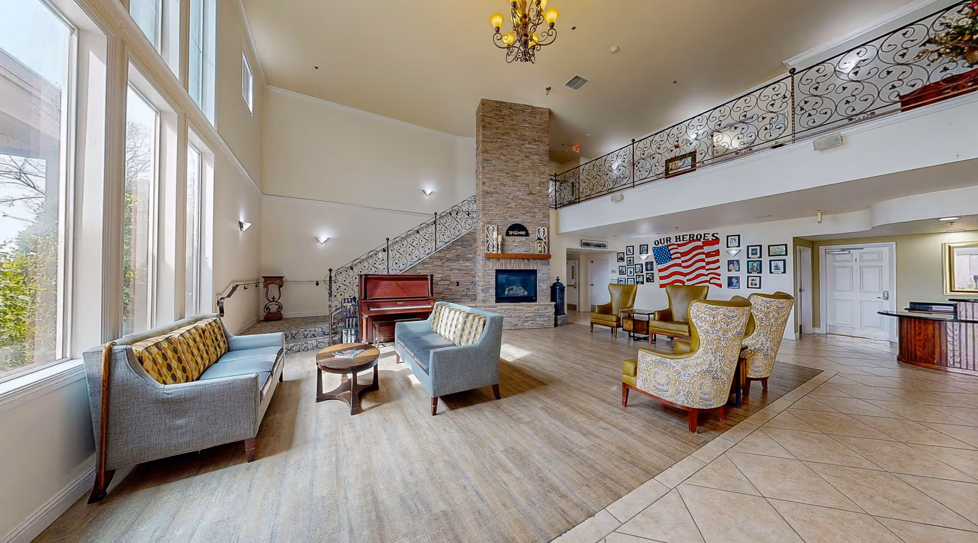 A spacious and well-lit living room area with large windows on the left side allowing natural light to fill the space. The room features two blue sofas with patterned cushions, a round wooden coffee table, and several upholstered armchairs arranged near a stone fireplace. A decorative wrought iron railing lines the upper balcony overlooking the room. On the far wall, there is a display with an American flag and framed photos under the words 'OUR HEROES'. A chandelier hangs from the ceiling, and a piano is positioned near the fireplace.