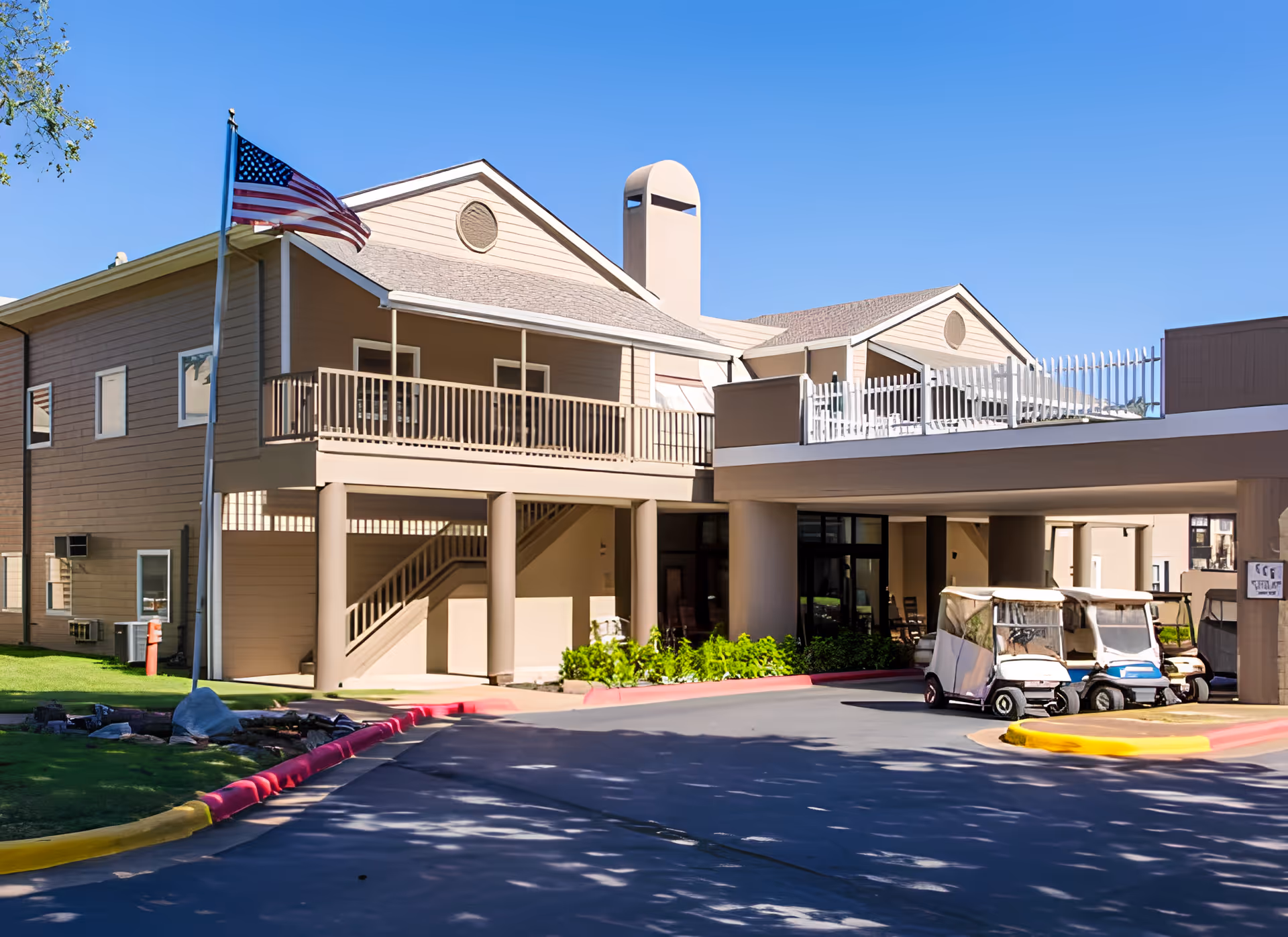 Exterior view of a two-story beige building with a covered entrance and a balcony. Several golf carts are parked near the entrance, and an American flag is flying on a flagpole to the left. The sky is clear and blue.