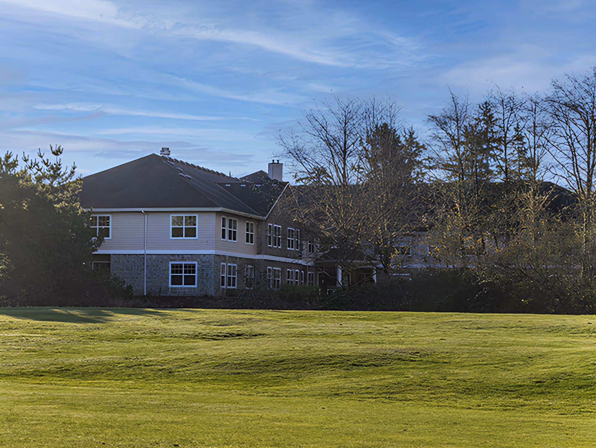 Exterior view of a two-story building with a dark roof and light-colored walls, surrounded by trees and a large grassy area in the foreground under a partly cloudy sky.