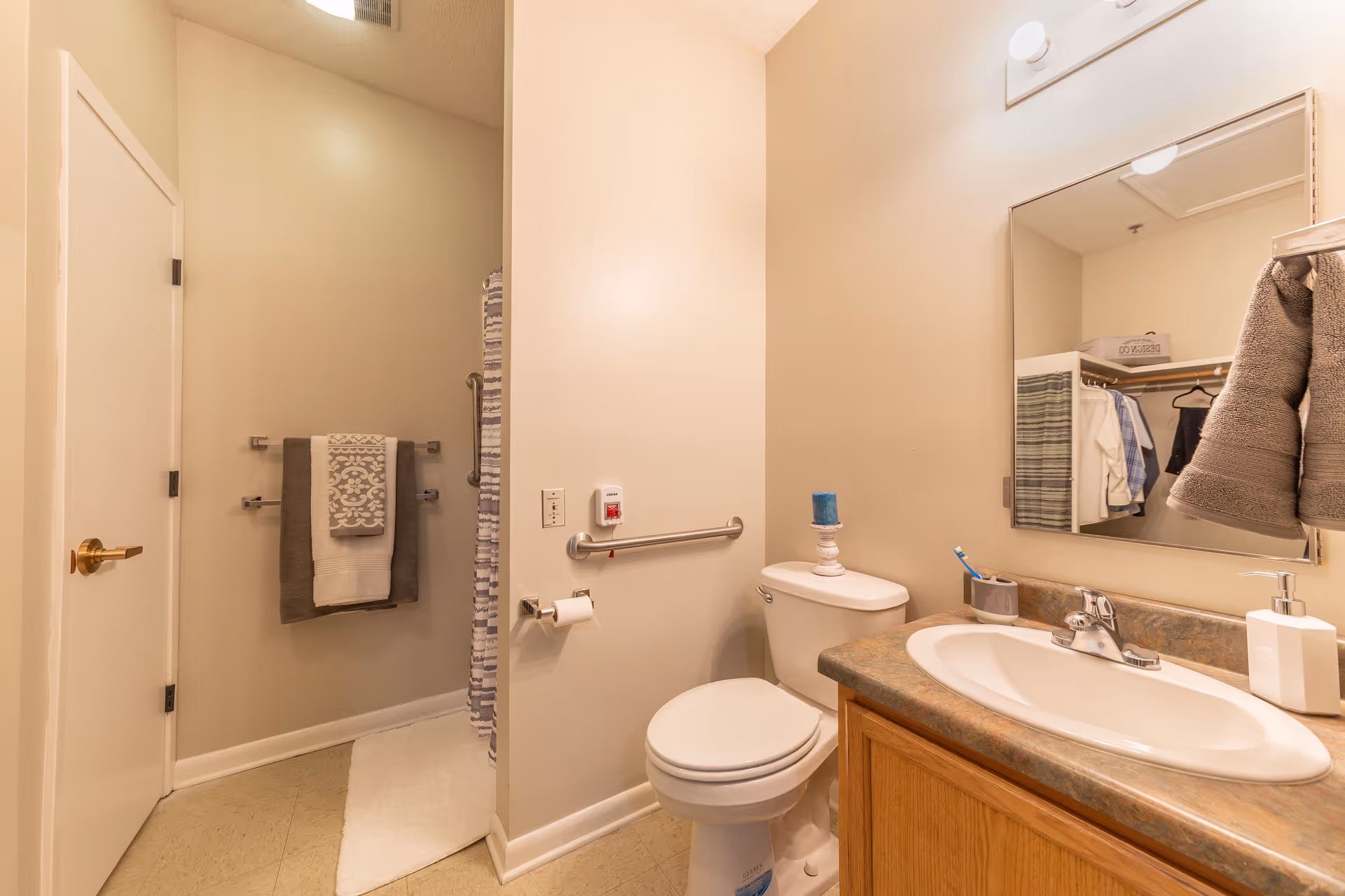 A clean and well-lit bathroom with a white toilet, a sink with a countertop, a mirror above the sink, a towel rack with folded towels, and a shower area with a striped shower curtain. The walls are painted beige, and there is a grab bar next to the toilet for accessibility.