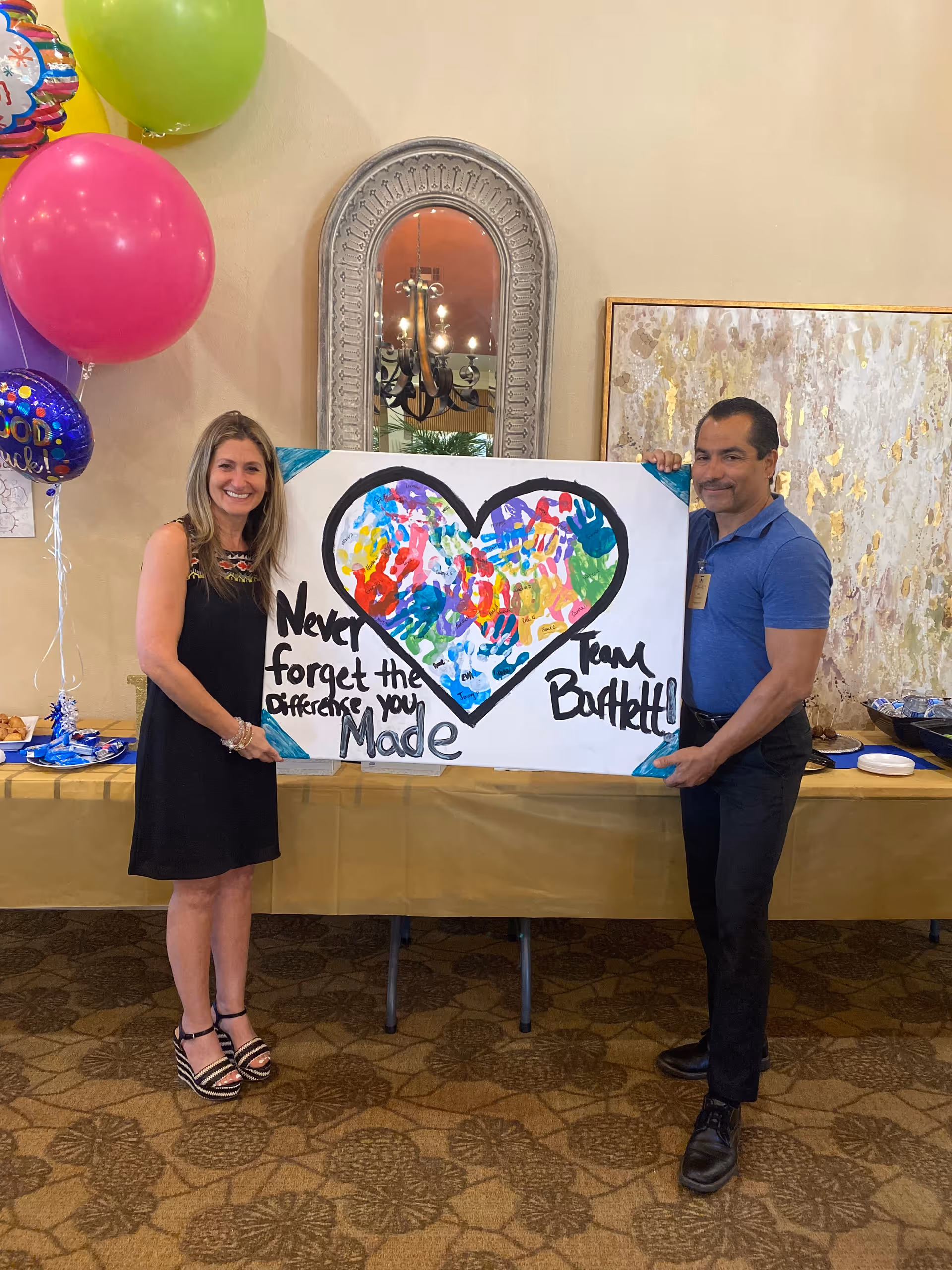 Two people standing indoors holding a large poster with a heart filled with colorful handprints. The poster reads 'Never forget the difference you made Team Bartlett'. There are balloons on the left side and a table with food and plates behind them.