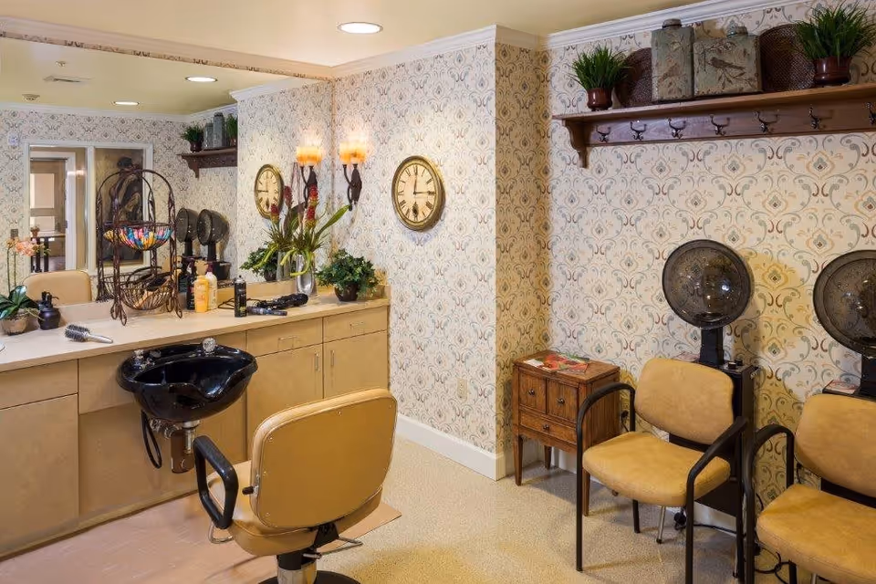 Interior of a hair salon area with a styling chair in front of a counter with a black sink, hair care products, and a large mirror. The walls have patterned wallpaper with two round clocks and wall sconces. There are two vintage hair dryers with chairs underneath and a small wooden side table with magazines. A shelf above holds decorative boxes and plants.