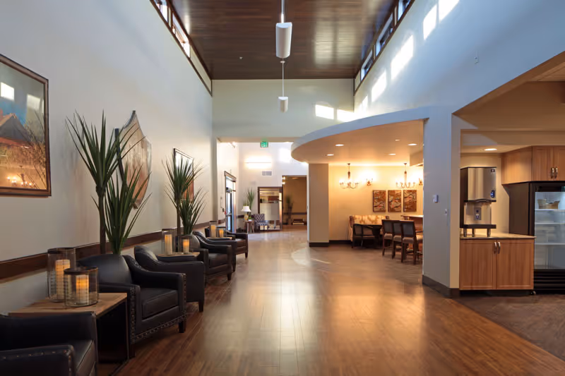 A spacious, well-lit interior hallway with wooden flooring and a high ceiling. On the left side, there are several black leather armchairs with side tables holding decorative candles and tall green plants. The right side features a curved wall enclosing a dining area with a chandelier and framed artwork on the wall. Further back, there is a kitchenette area with wooden cabinets and a refrigerator. Natural light streams in through high windows on the right wall.