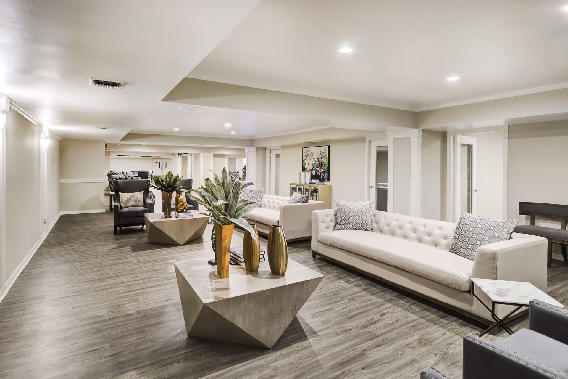 Spacious modern lounge with light-colored sofas, geometric coffee tables, plants, and decorative vases.