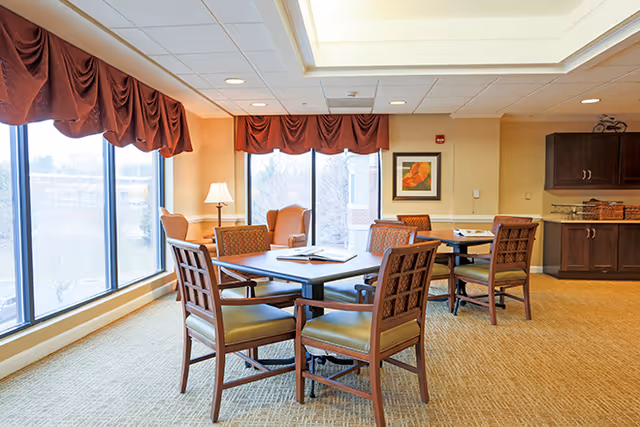 Communal dining area with wooden tables and chairs near large windows and a small kitchenette.