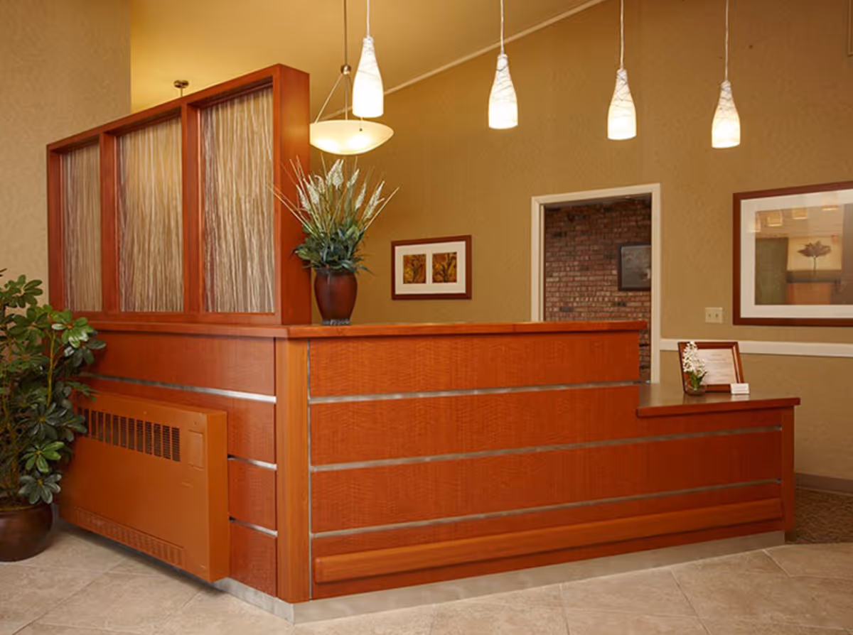 Reception area with a wooden front desk featuring a potted plant on top, pendant lights hanging from the ceiling, framed artwork on the walls, and a potted plant on the floor to the left.
