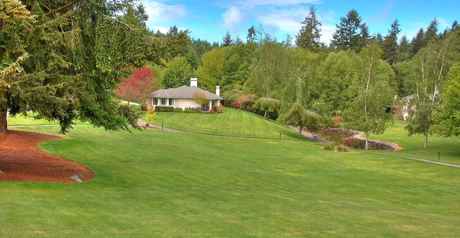 A large, well-maintained grassy lawn with a paved walkway winding through it, surrounded by various trees and shrubs. In the background, there is a single-story building with a gray roof and white exterior, set against a backdrop of dense forest and a partly cloudy blue sky.