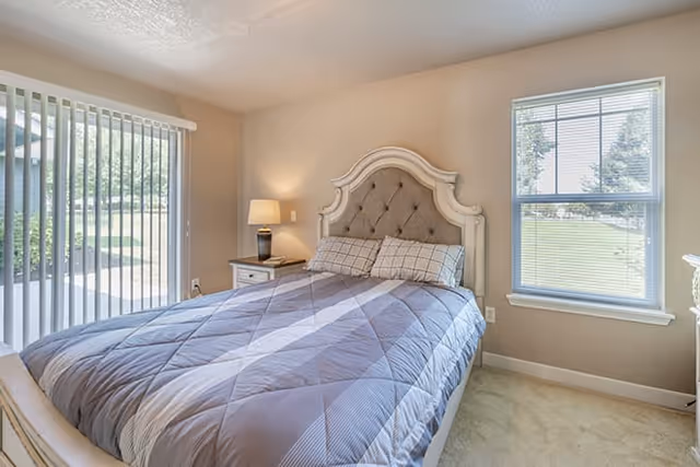 A bright bedroom with a large bed featuring a tufted headboard and a gray quilted bedspread. There is a nightstand with a lamp on the left side of the bed, a window with blinds on the right wall, and sliding glass doors with vertical blinds on the left wall, allowing natural light to fill the room.