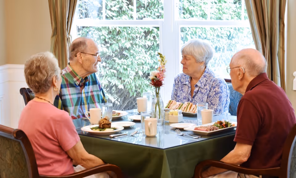 Four elderly residents sitting around a dining table by a window, eating and talking.