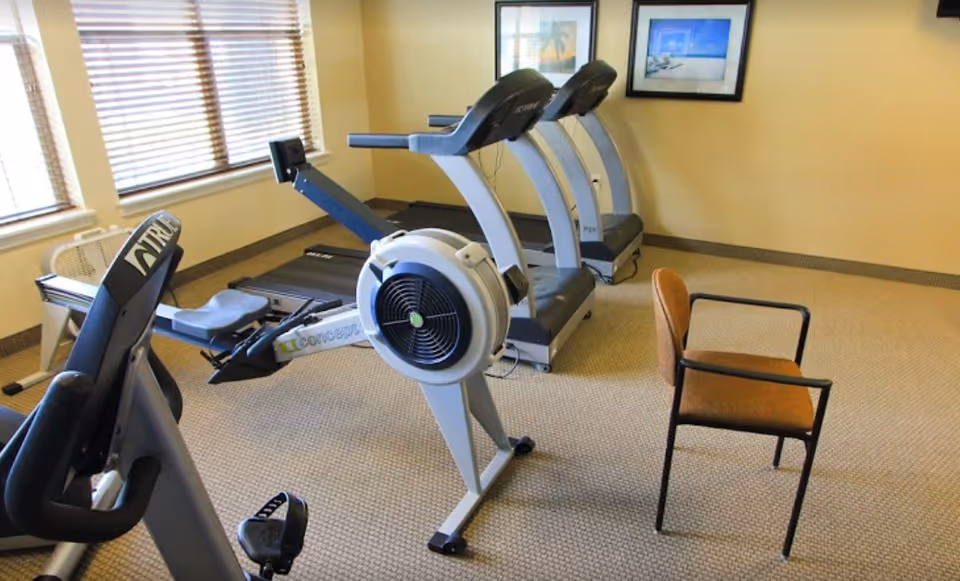 A small exercise room with two treadmills, a rowing machine, and an exercise bike. There is a single brown chair with black armrests in the room. The walls are light yellow with two framed pictures hanging, and there are windows with blinds letting in natural light.