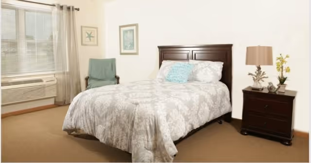 Bright bedroom with a made double bed featuring patterned bedding, a wooden headboard and nightstand with lamp, a chair, and a window with curtains.
