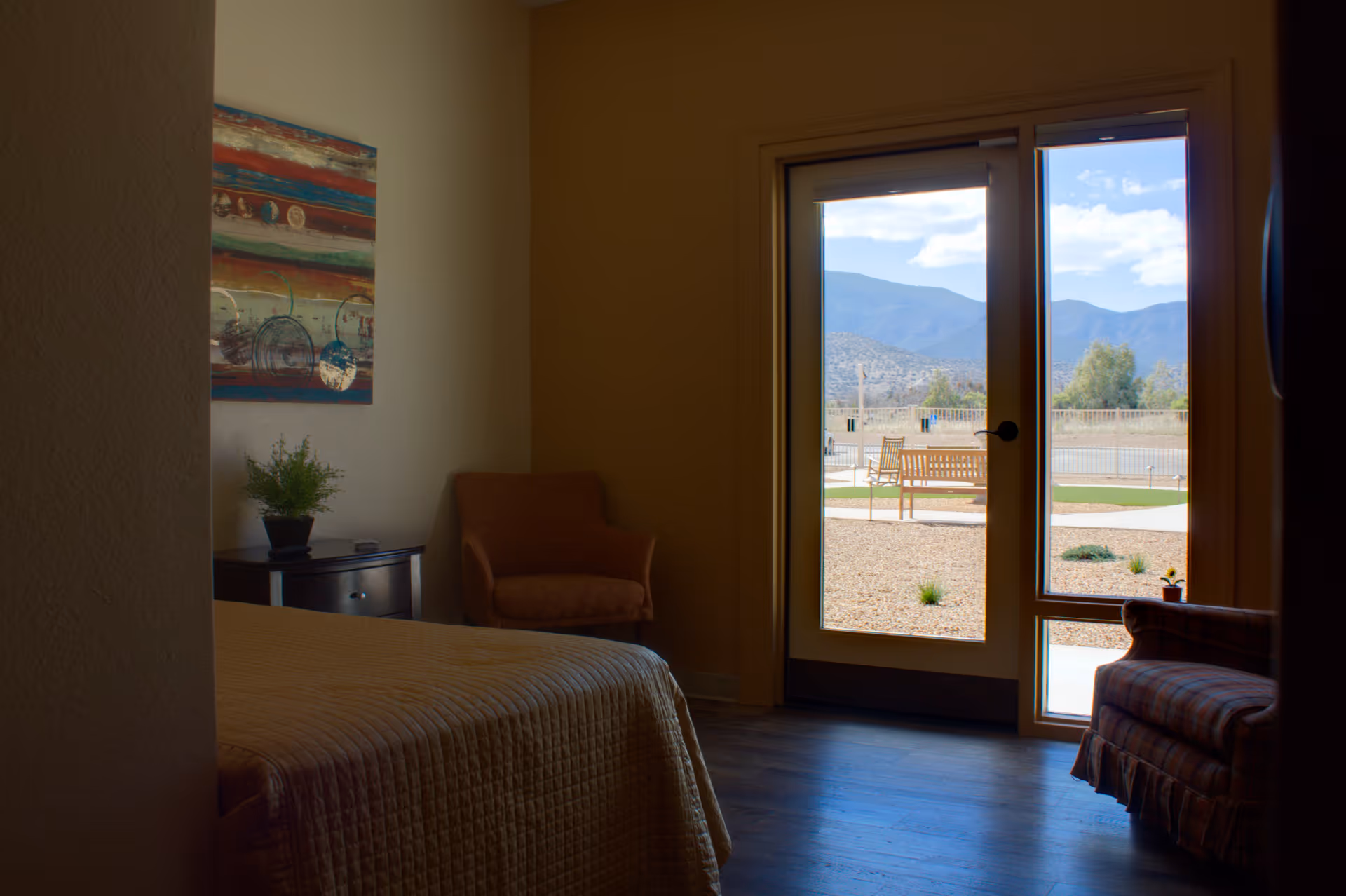 A bedroom in an assisted living facility with a bed covered in a beige quilt, a small dark wooden nightstand with a potted plant, an armchair, and a large glass door and window showing an outdoor area with benches and mountains in the background.