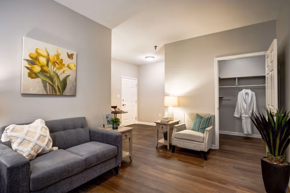 A cozy living area with a gray sofa adorned with a white textured pillow, a beige armchair with a green striped cushion, and two wooden side tables. One side table has a lamp and books, while the other has a small plant and a framed photo. A painting of yellow tulips with a butterfly hangs on the wall above the sofa. The room has wooden flooring and light gray walls. In the background, there is an open closet with a white robe hanging inside and a white door leading to another area.