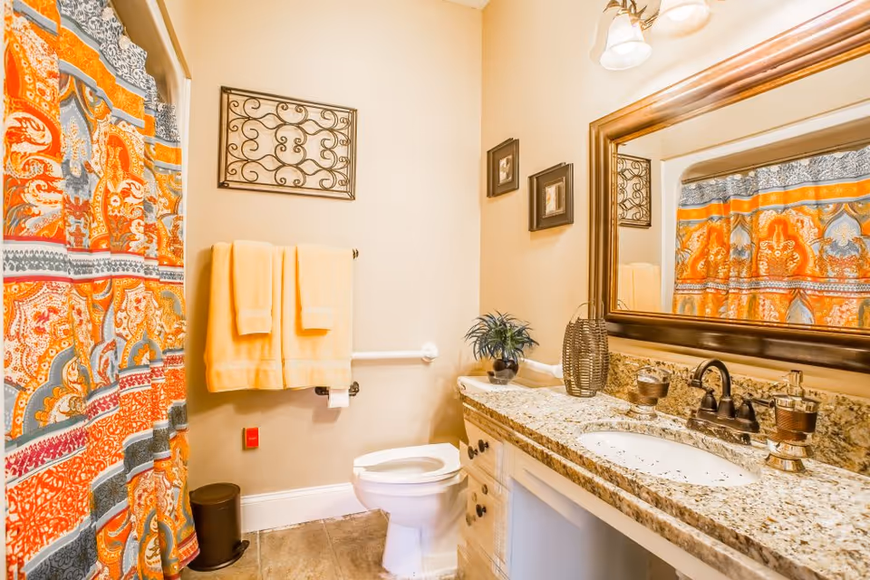 A bright bathroom with a colorful orange, blue, and white patterned shower curtain. There are two yellow towels hanging on a towel rack above a toilet. The bathroom has a granite countertop with a sink, a large framed mirror, and decorative items including a small plant and a woven vase. The walls are painted beige and there are two small framed pictures hanging next to the mirror.