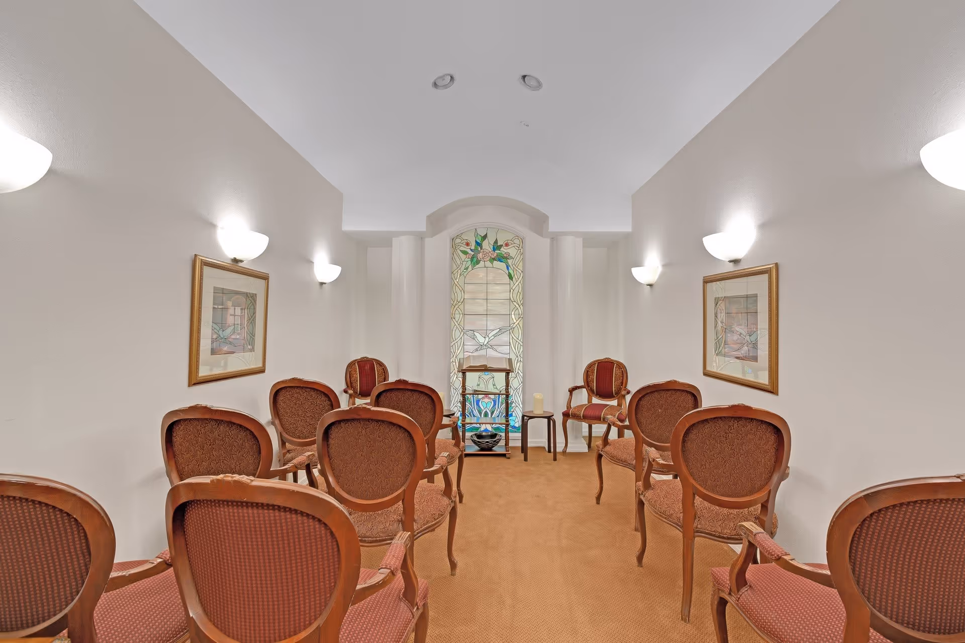 A small chapel or meditation room with rows of wooden chairs upholstered in red fabric facing a stained glass window with floral and bird designs. The room has beige walls, wall-mounted lights, framed artwork on both sides, and a small table with a candle and a bowl near the window.