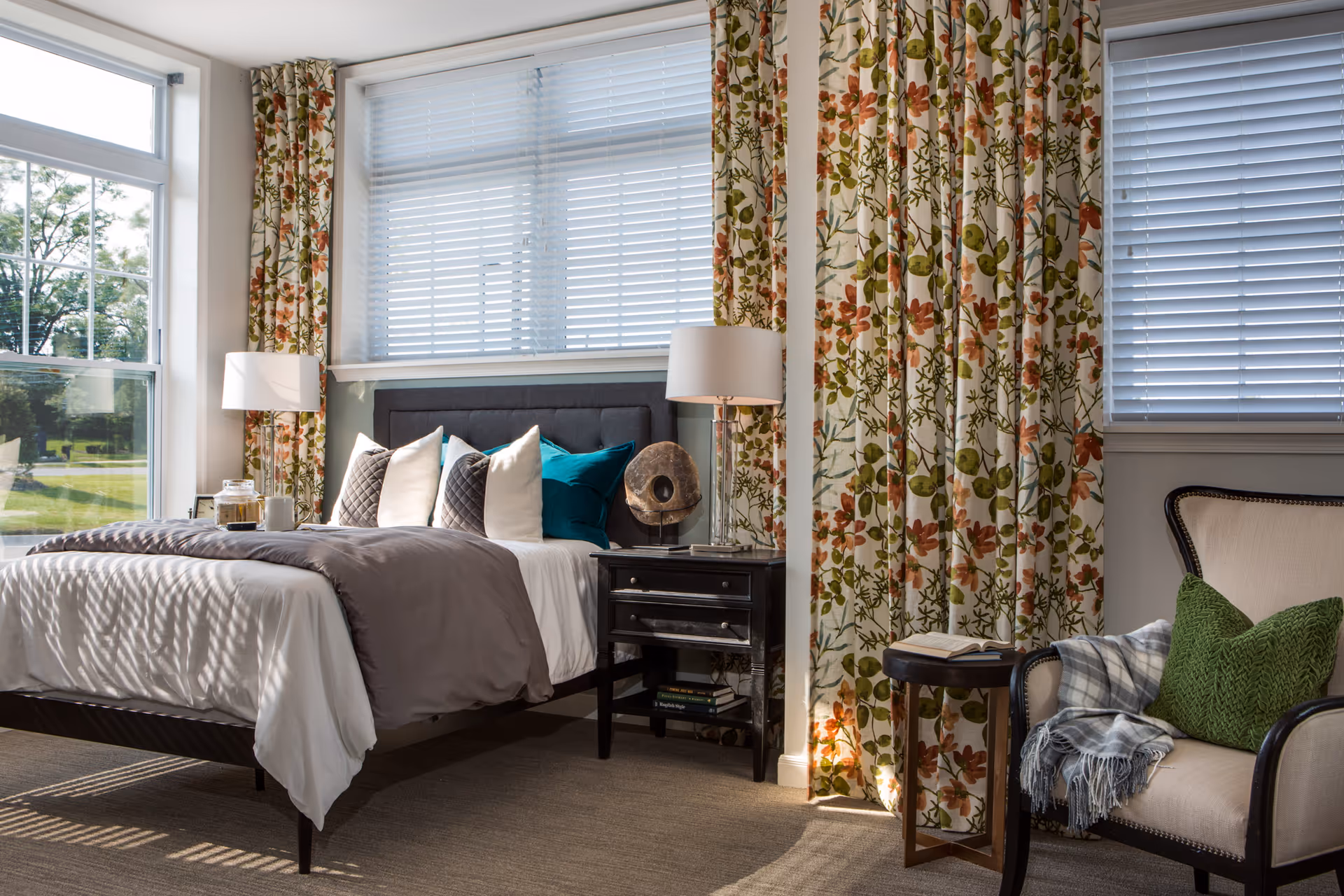A bright and cozy bedroom featuring a neatly made bed with white and gray bedding and multiple pillows. The room has large windows with floral curtains and white blinds, allowing natural light to fill the space. Beside the bed is a dark wooden nightstand with a lamp and decorative item. There is also a comfortable armchair with a green pillow and a plaid throw blanket, next to a small round side table with an open book.